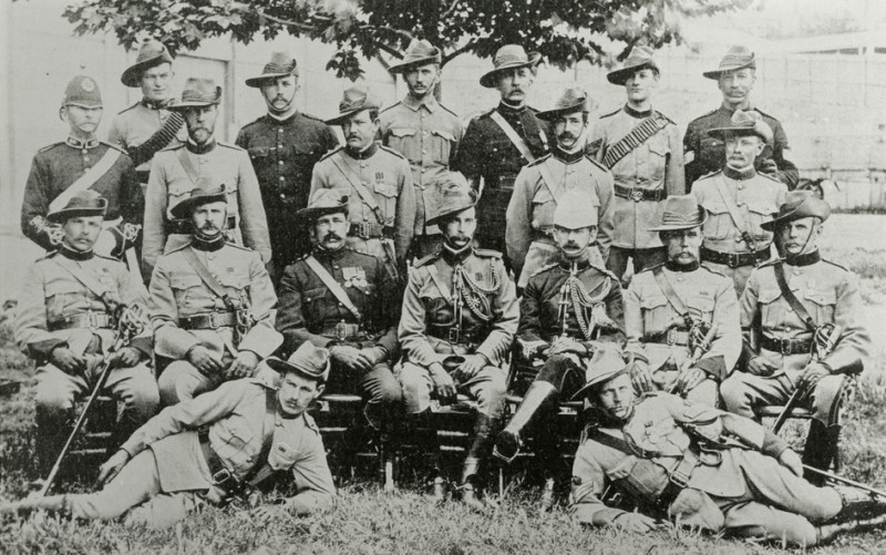 20 men pose in rows for a photograph. They wear military uniforms including hats, belts, boots and buttoned-up jackets.