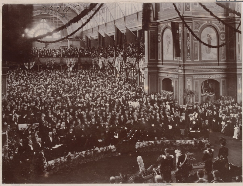 A large crowd stands inside a large building. They face towards a platform where several people stand in formal clothing. The inside of the building is decorated with flags and streamers.