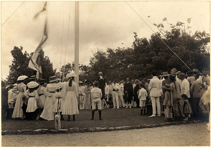 Sepia photo showing a crowd of women, men and children standing around a large flag pole. A man in a black robe and barrister's wig stands on a stepladder in the centre of the crowd. Most of the crowd are wearing white clothing and hats.