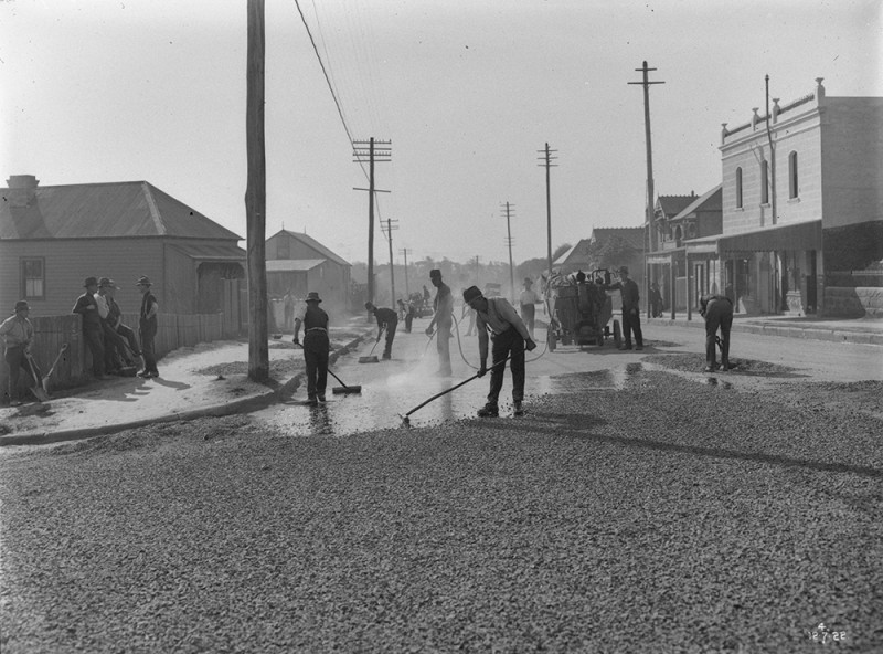 Black and white photo of seven men washing and sweeping a main road with shops and electricity poles in the background. 