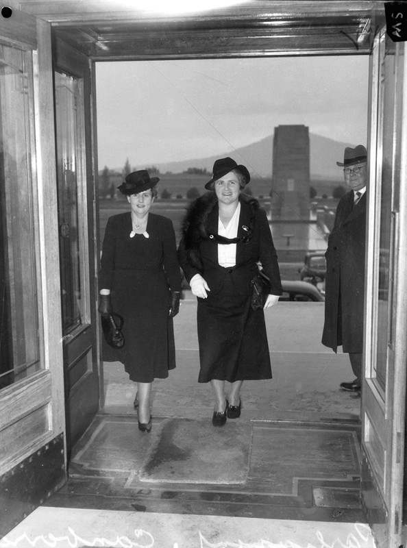 A black and white photo of two women wearing dresses, hats, gloves and carrying purses. The photo is taken from inside a building as they stand and walk together into it.