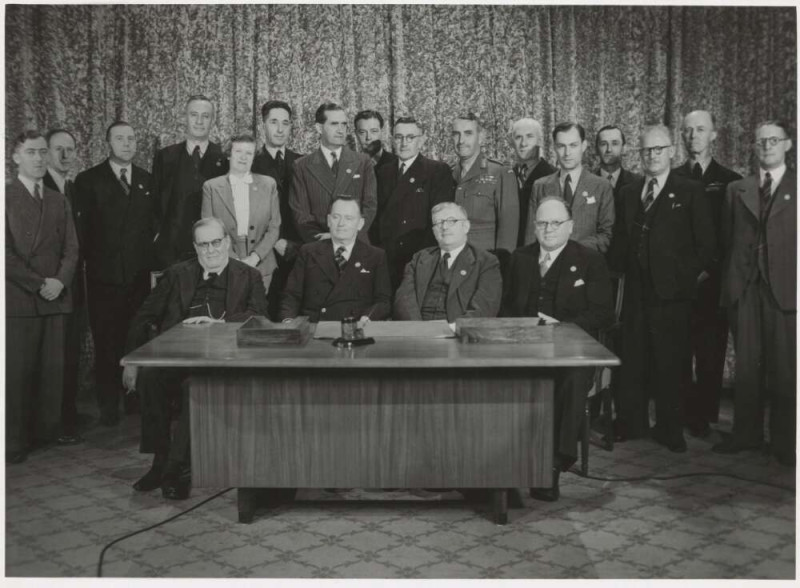 19 men and 1 women all dressed in suits appear in a black and white photo facing towards the camera. 4 of the men sit at a desk. Everyone else stands in rows behind them. 