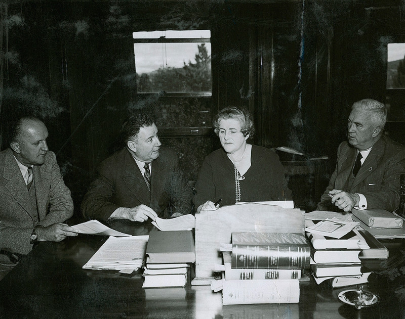 A black and white photo of a woman and three men sitting to work at a desk. The desk is covered with several books, papers and an ash tray.