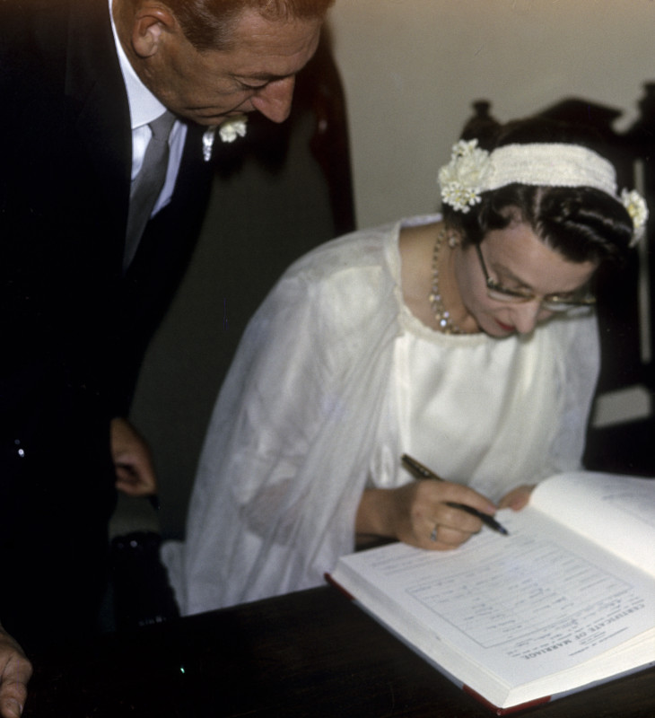 Woman in white dress sitting, signing register with man in suit standing behind looking over her shoulder.