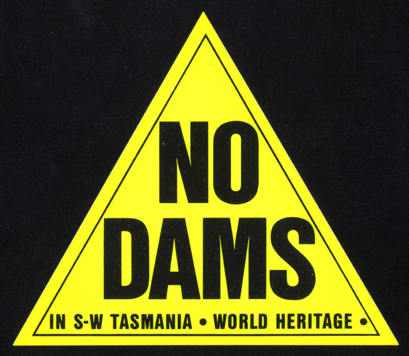 A yellow triangular sticker with the text ‘NO DAMS IN S-W TASMANIA WORLD HERITAGE’ printed on it in black. 