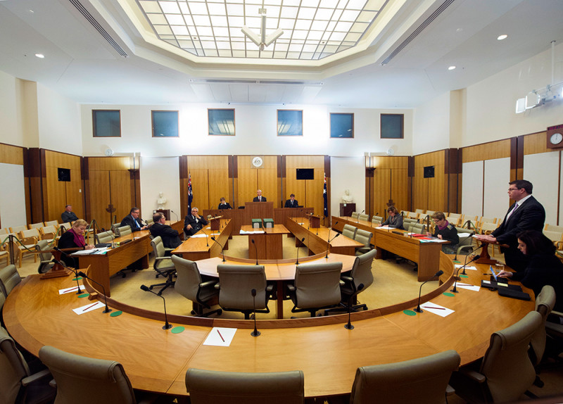 A wood-panelled room with tables and chairs arranged in a U-shape. A man stands up to give a speech while other people sit.