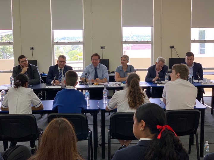 Six adults sit at a table facing 4 secondary school students seated at another table. The adults are listening to the students.