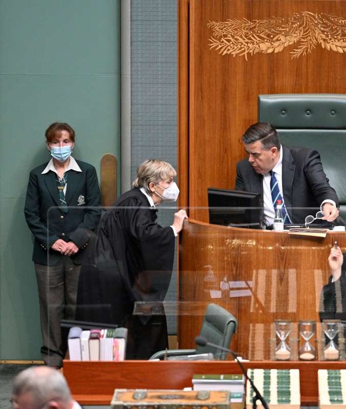 The Clerk, wearing a black gown, leans in to speak to the Speaker sitting in a raised seat surrounded by wood.