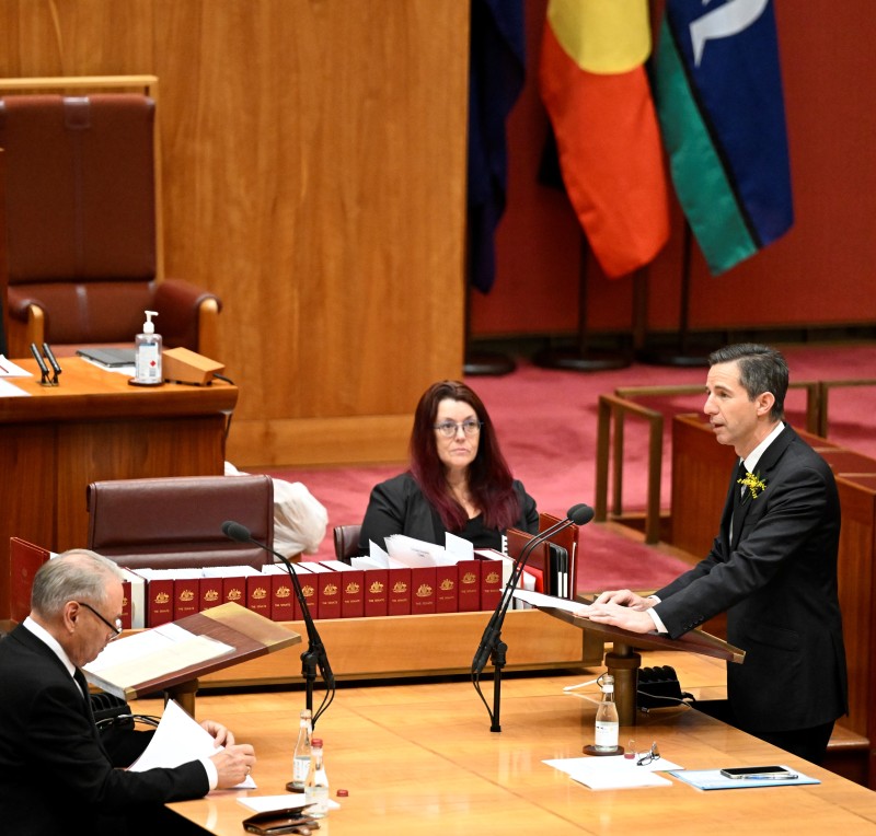 A man in a suit stands at a table and is speaking in a red room. A woman and a man in dark suits also sit at the table.