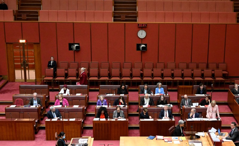 In a red room, men and women sit on 2 seater benches with desks in front of them. Behind them are rows 2 rows of chairs.