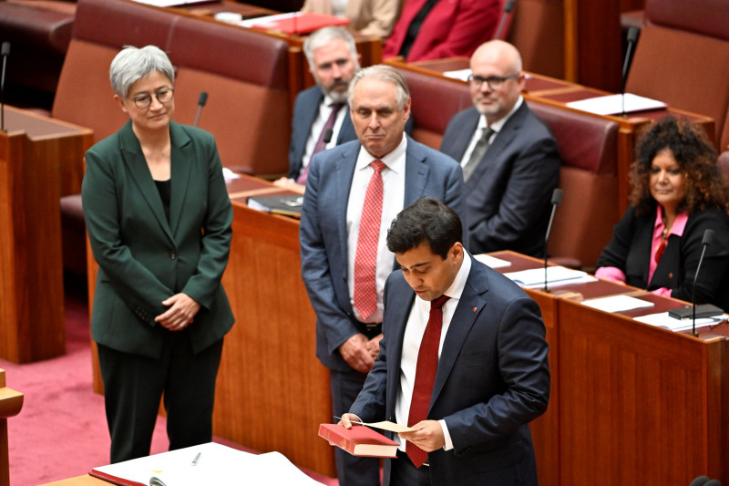 A new senator stands in the Senate. He holds a holy book and reads an oath of allegiance.